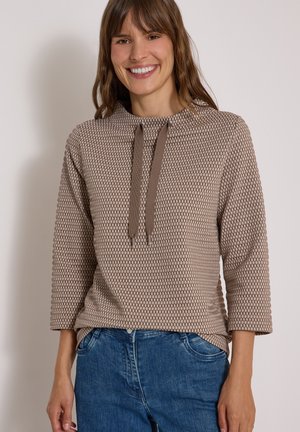 Kvinde smilende iført en beige langærmet top med mønster og snørredetalje samt blå jeans, stående mod en ensartet lys baggrund.