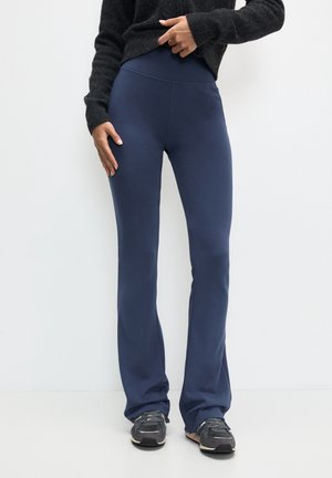 Person trägt dunkelblaue, eng anliegende Schlaghosen, schwarze Sneaker und einen schwarzen Langarm-Pullover und steht vor einer schlichten weißen Wand.