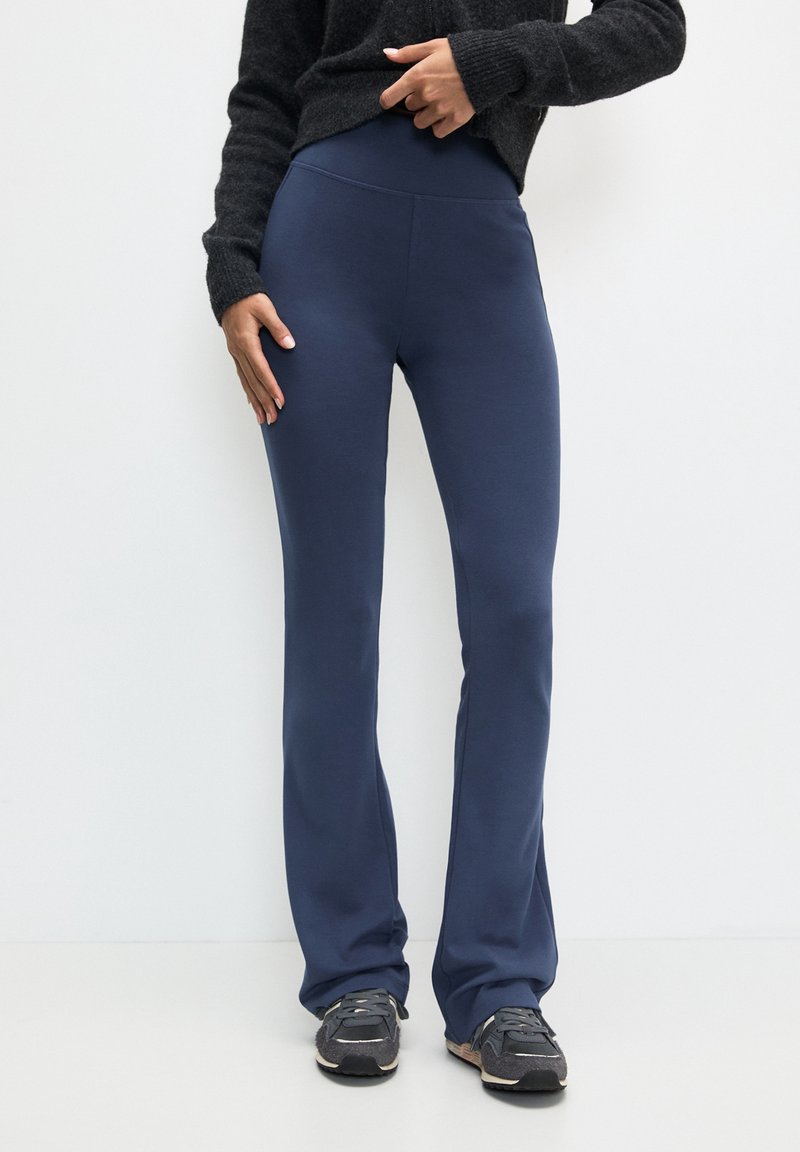 Personne portant un pantalon évasé ajusté bleu marine, des baskets noires et un pull à manches longues noir, debout contre un mur blanc uni.