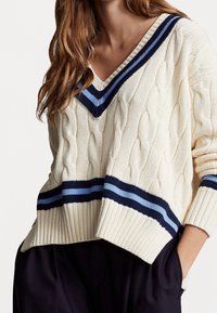 Krämfärgad kabelstickad tröja med V-ringning, med marinblå och ljusblå randiga detaljer vid halsringningen och fållen. Avslappnad passform, ribbade ärmslut.