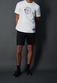 T-shirt blanche à manches courtes avec logo circulaire, associée à un short noir. Des baskets noires et des chaussettes complètent le look. Design simple et décontracté.