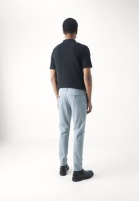 Camisa polo negra de manga corta con tejido texturizado, combinada con pantalones grises claros a medida y botas negras hasta el tobillo, vista desde atrás.