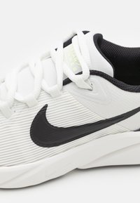 Chaussure de sport blanche avec texture rayée, logo noir Swoosh de Nike, col contrastant noir et semelle blanche plate. Les lacets sont bien visibles.