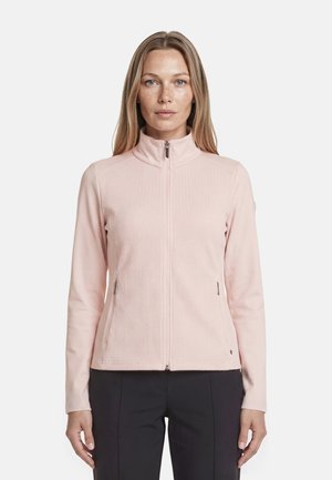 Mujer con chaqueta rosa claro con cremallera y cuello alto, y pantalones negros, de pie mirando hacia adelante contra un fondo liso.