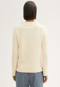 Pull en crème avec une texture côtelée, manches longues et un col montant, associé à un pantalon bleu clair bien ajusté.