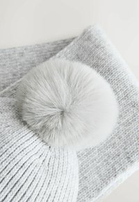 Pelēks adīts cepure ar lielu kažokādas pom-pomu augšpusē, parādīts kopā ar atbilstošu pelēku adītu šalli, kurai ir faktūras raksts.