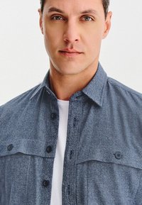 Mężczyzna o zielonych oczach, ubrany w niebieską koszulę zapinaną na guziki na białą koszulkę, patrzący na wprost z neutralnym wyrazem twarzy na jednolitym tle.