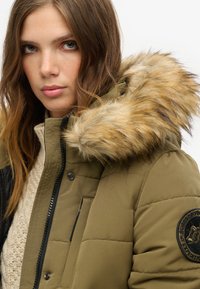 Poduszka oliwkowa kurtka z kapturem z sztucznego futra, czarnym zamkiem błyskawicznym i naszywką na rękawie. Wnętrze wykończone jest teksturowanym, kremowym swetrem.