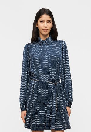 Ženska v mornarsko modri obleki z dolgimi rokavi z belimi pikami, ovratnikom in verižnim pasom, stoji ob enostavni svetli podlagi.