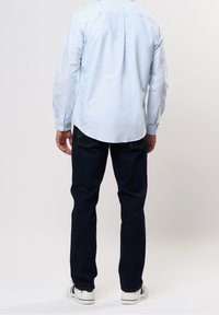 Camisa de manga larga de color azul claro con botones y dobladillo redondeado, combinada con jeans de mezclilla oscura y zapatillas blancas con detalles en azul y rojo.