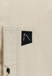 Tessuto in tela bianco sporco, con una patch quadrata nera che presenta un design astratto bianco e un dettaglio con bottone argentato sul lato.