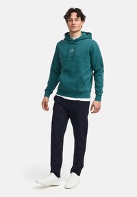 Sudadera de algodón en color verde azulado con un bolsillo frontal y cordones, combinada con pantalones oscuros y zapatillas blancas. El modelo está de pie frente a un fondo liso.