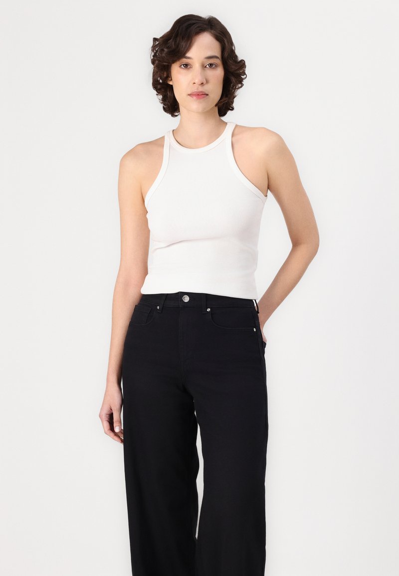 Camiseta de tirantes blanca con un cuello alto, combinada con unos pantalones vaqueros de pierna ancha negros a la cintura. La tela parece suave con un detalle mínimo.