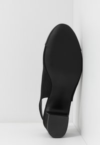 Semelle d'une chaussure à talon en daim noir avec un talon en caoutchouc texturé, positionnée verticalement contre un mur d'angle blanc et le sol.