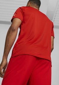 Červené sportovní tričko s krátkými rukávy, texturovaná tkanina, s logem na rameni. Doprovázeno odpovídajícími červenými šortkami.