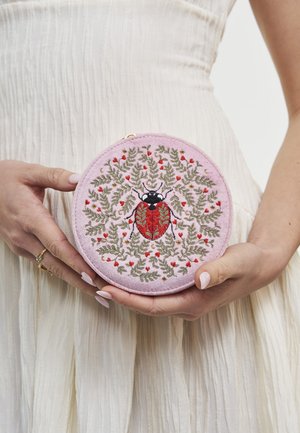 Mani che tengono una pochette rotonda rosa con una coccinella ricamata in rosso e nero, circondata da foglie verdi e piccoli fiori rossi.