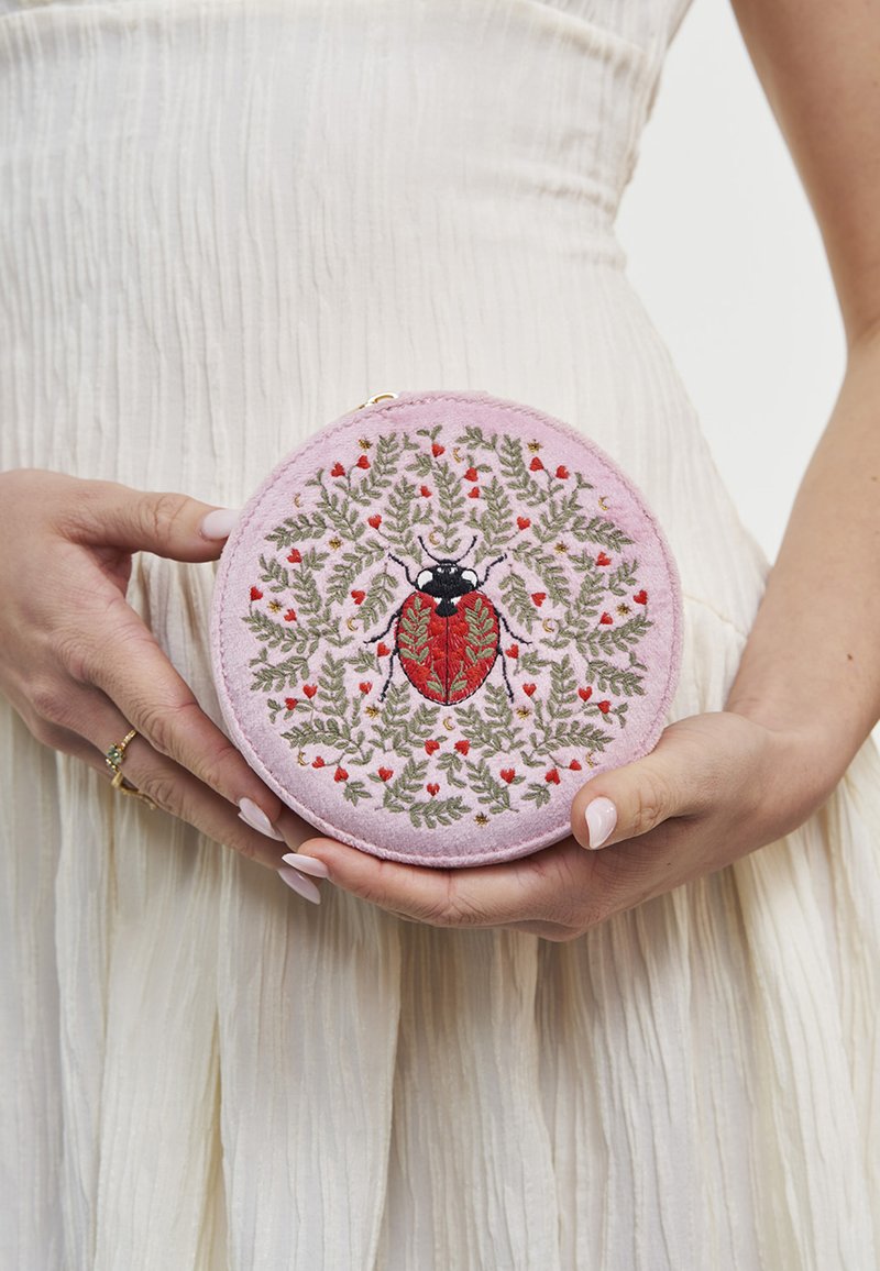 Ruky držia okrúhle ružové vrecúško s vyšitou červenou a čiernou lienkou obklopenou zelenými listami a malými červenými kvetmi.