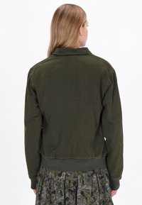 Oliwkowa kurtka z kołnierzem i ściągaczem. Posiada teksturowany materiał, długie rękawy i luźny krój. Widok z tyłu, brak widocznych elementów metalowych.