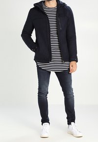 Giacca cappuccio navy con zip frontale, tasche sul petto e tessuto texturizzato. Abbinata a una maglietta a maniche lunghe a righe e jeans scuri. Sneakers bianche.