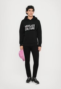 Čierna mikina s kapucňou a bielym logom "Versace Jeans Couture", skombinovaná s čiernymi priliehavými nohavicami. Drží ružovú čiapku s textom. Čierne tenisky s akcentmi.