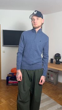 Kék kötött pulóver cipzáras nyakkal, laza szabású olívazöld nadrággal párosítva. Kéz tetoválásokkal és sötét sapkával.