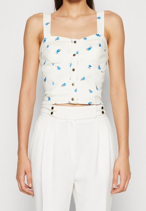 Kobieta ubrana w biały krótki top na guziki z niebieskim kwiatowym wzorem oraz białe spodnie z wysokim stanem, z guzikami przy talii.