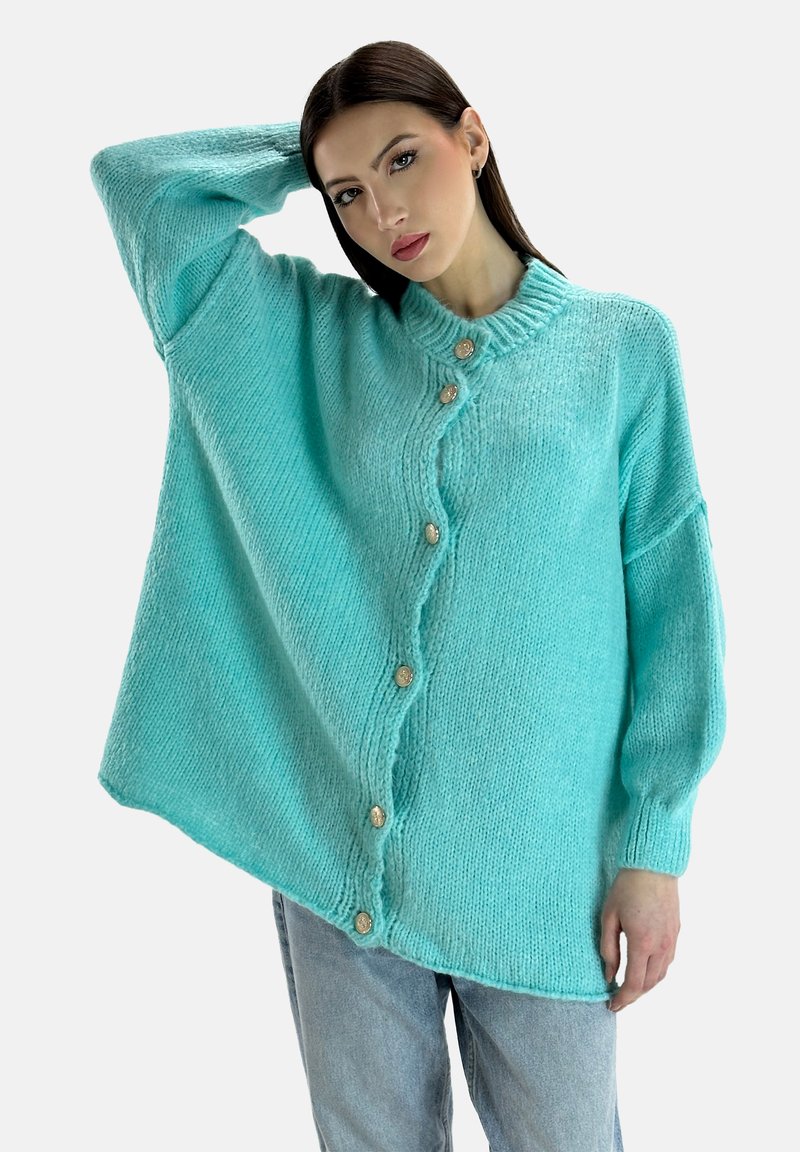 Türkise Strickjacke mit hohem Kragen, großen Knöpfen und übergroßen Ärmeln, kombiniert mit hellen Jeans.