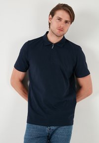Námořnická modrá polokošile s krátkým rukávem a zipovým límcem, vyrobená z hladkého materiálu, s přiléhavým střihem a volným stylem. Nosená s modrými džíny.
