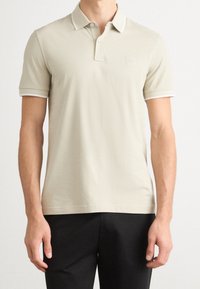 Polo beige clair avec un col côtelé et des bordures blanches, présentant un petit logo sur la poitrine et des manches courtes. Design à coupe régulière.