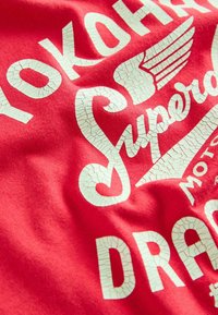 Röd bomulls T-shirt med stor sprucken vit text med orden "Yokohama Superdry" och dekorativa vingaccent.