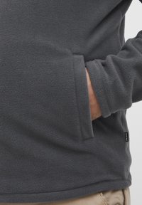 Sweat-shirt en molleton gris avec une texture douce, manches longues et poches latérales. Dispose d'une étiquette de marque visible en bas à gauche.