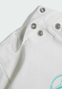 Angolo di un indumento in cotone bianco con chiusure a pressione in metallo. Presenta una trama liscia e un dettaglio del logo stampato in azzurro.