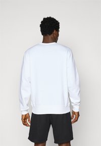 Sweatshirt blanc avec des poignets et un ourlet côtelés, design à col rond, texture lisse. Porté avec un short noir, montrant une coupe décontractée vu de l'arrière.