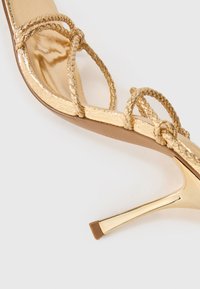 Sandalía dorada de tacón alto con correas trenzadas, un acabado metálico brillante y un tacón de aguja afilado. Los acentos texturizados realzan el diseño.