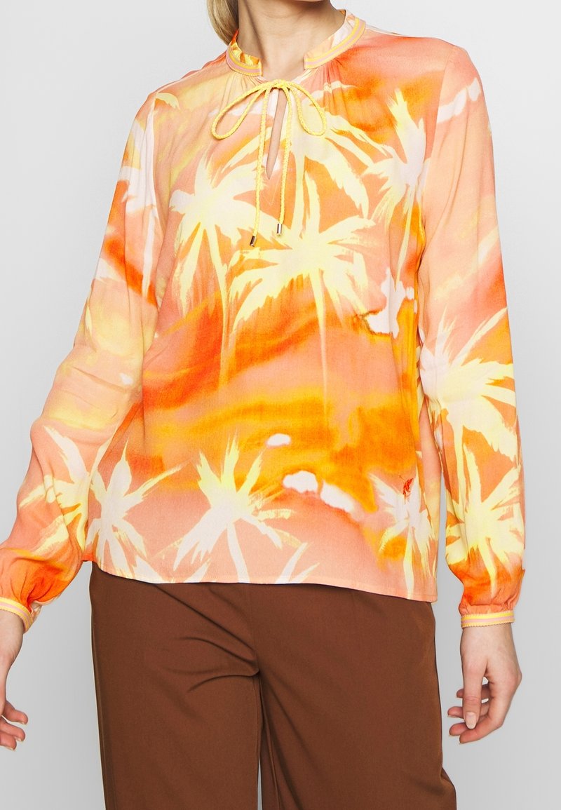 Femme portant un chemisier à manches longues à imprimé tropical orange et jaune avec un col à nouer, assorti à un pantalon marron.