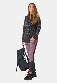 Czarna i biała kurtka z zamkiem błyskawicznym i wysokim kołnierzem, w połączeniu z fioletowymi i czarnymi spodniami, niosąca czarny plecak, w czarnych butach.