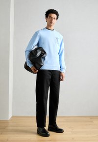 Light blue sweatshirt with logo, black trousers, and black shoes. Holds a large, crumpled black bag. Simple design, smooth texture.