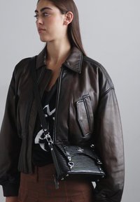 Giacca bomber in pelle marrone con texture trapuntata, abbinata a una piccola borsa a tracolla nera con motivo coccodrillo e hardware in argento.