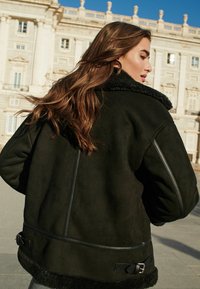 Svart faux shearling-jacka med en bred krage, sidodragkedjedetaljer och spännen vid fållen. Mjuk textur och oversized passform.