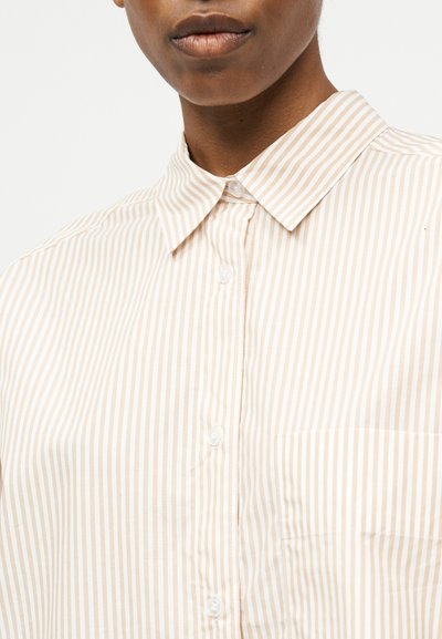 Κοντινό πλάνο ενός ατόμου που φοράει πουκάμισο με κάθετες ρίγες σε μπεζ και λευκό χρώμα, με κουμπιά, μυτερό γιακά και τσέπη στο στήθος.