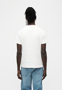 Λευκό βαμβακερό μπλουζάκι με κοντά μανίκια, στρογγυλή λαιμόκοψη και λεία υφή, φορεμένο με ανοιχτό μπλε τζιν παντελόνι. Απλή όψη από πίσω.