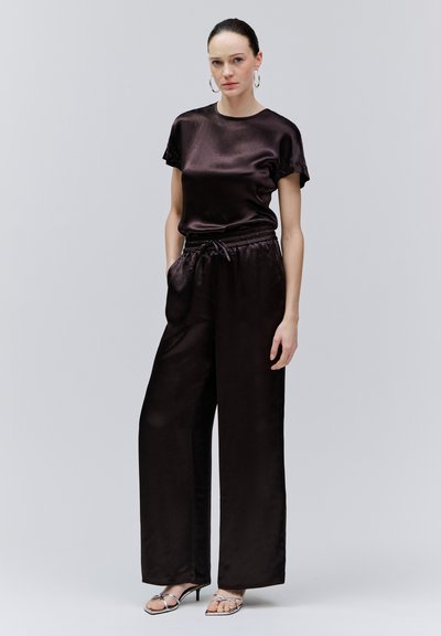 Femme debout portant un haut noir en satin à manches courtes et un pantalon à jambes larges, avec des boucles d'oreilles argentées en forme d'anneau et des sandales à talons à brides, sur un fond gris.