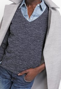 Suéter de punto navy con escote en V, sobre una camisa de denim azul claro, complementado con un abrigo gris claro. Tejido texturizado, diseño ajustado.