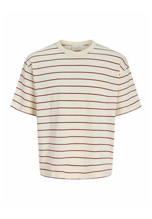 Băiat cu păr blond scurt stă privind spre dreapta, purtând o cămașă crem cu dungi roșii, pantaloni largi maro și adidași bej pe un fundal alb.