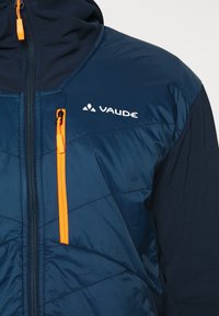 Marinblå isolerad jacka med quiltad textur, kontrasterande orange dragkedja och Vaude-logotyp på bröstet. Innehåller en huva och en ficka med dragkedja.