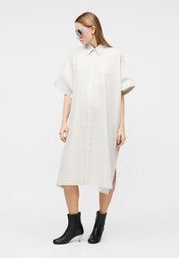 Frau trägt ein übergroßes weißes Hemdkleid, transparente Visier-Sonnenbrille und schwarze Stiefeletten mit dünnen Absätzen, steht vor einem einfarbigen Hintergrund.