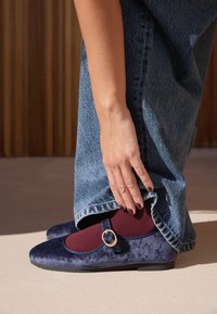 Granatowe baleriny wykonane z zamszu, z bordową elastyczną wstawką i złotym detalem klamry. Brzeg z denimu częściowo zakrywa buty.