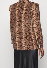 Blazer à motif peau de serpent marron en tissu texturé, avec une fente unique à l'arrière et trois boutons en décoration aux poignets.