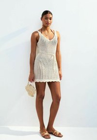 Gebreid, crème-kleurig jurk met een V-hals, een ceintuur op de taille en een scalloped zoom. Gecombineerd met bruine instappers en een kleine geweven handtas.