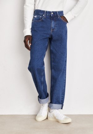 Person trägt hochgekrempelte blaue Jeans, weißes Langarmshirt und weiße Sneakers und steht an einer schlichten Wand.